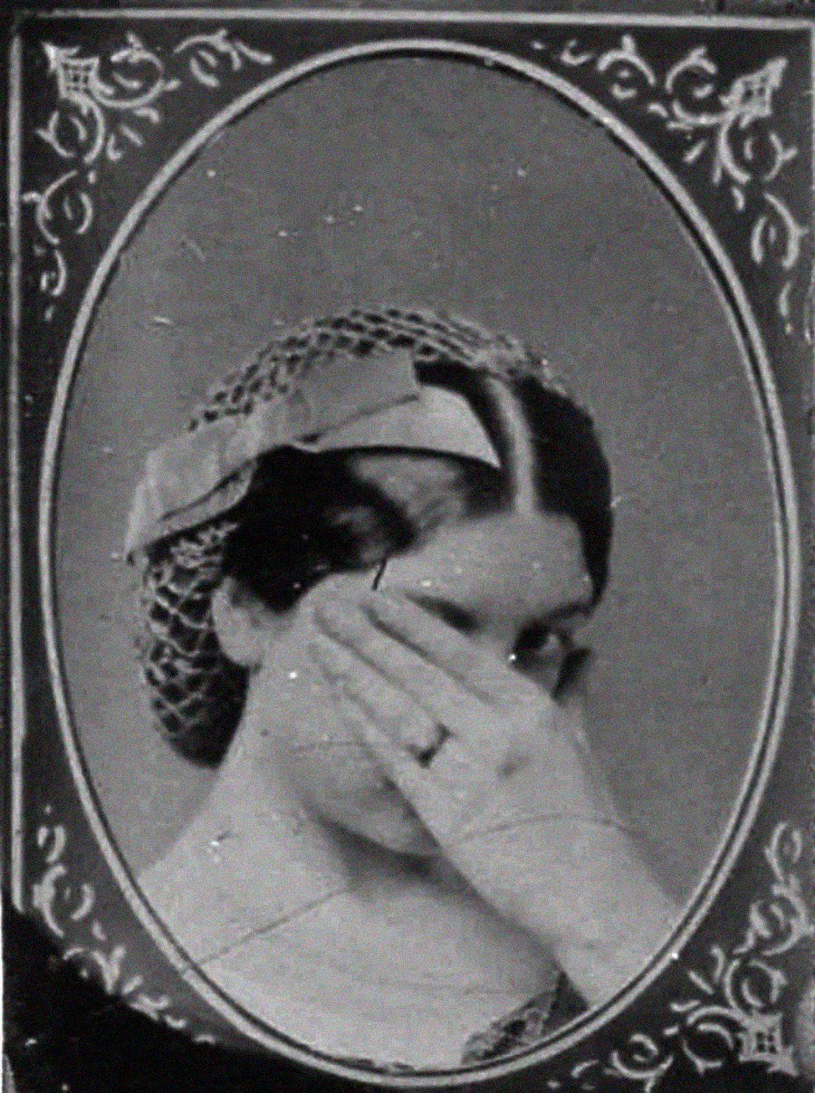 A vintage black-and-white portrait of a woman covering most of her face with her hand. She wears a hair net with a ribbon, and a ring is visible on her finger. Ornate patterns frame the oval photograph.