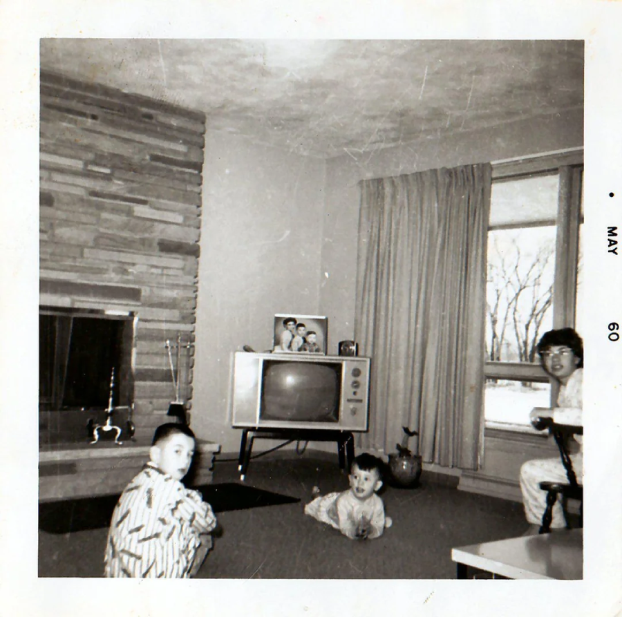 Black-and-white photo from May 1960 showing two children and an adult in pajamas in a living room with a TV, fireplace, large window with curtains, and family photos displayed on the TV.