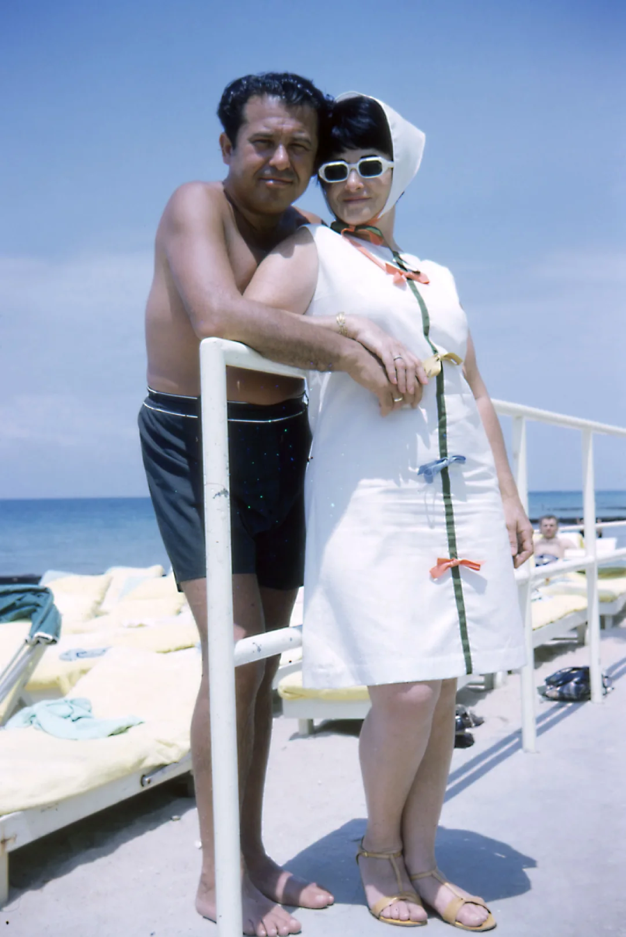 A man in swim trunks and a woman in a white dress, sunglasses, and headscarf pose by a metal railing on a sunny beach, with lounge chairs and the ocean in the background.