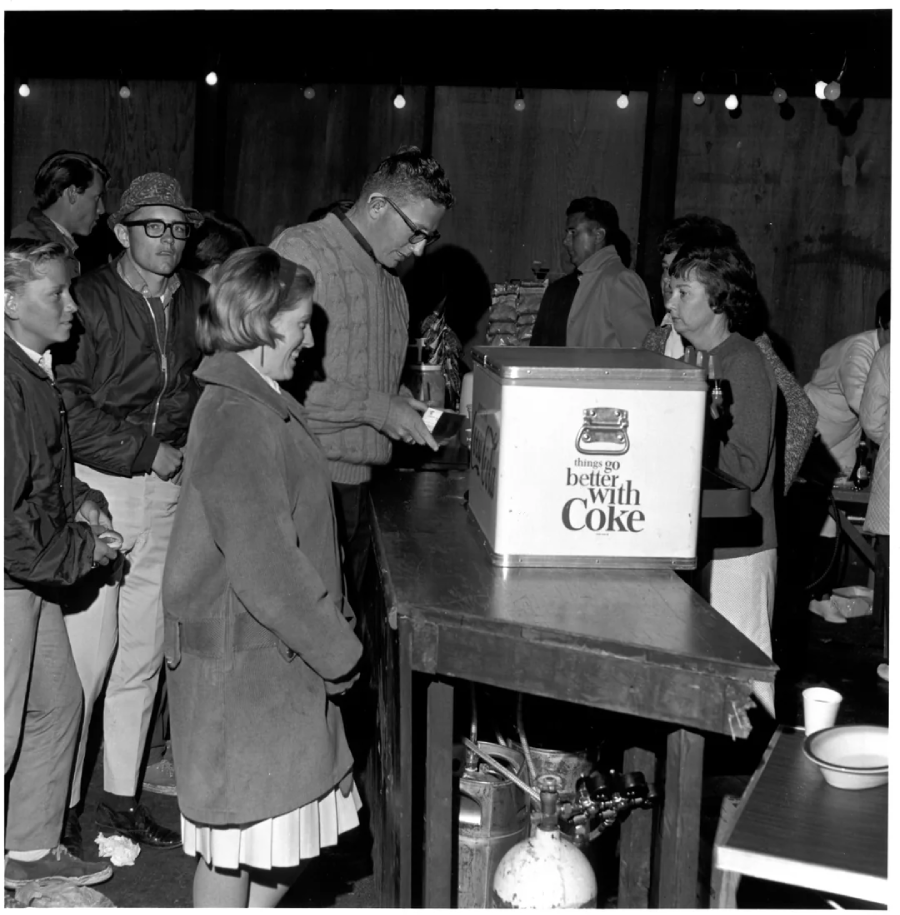 A group of people stand in line at an outdoor refreshment stand, where a man serves drinks from a cooler with a "things go better with Coke" sign. String lights hang above, and most people wear jackets.
