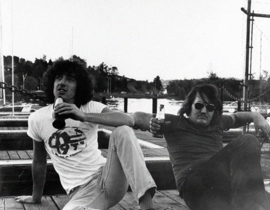 Two men relax on a wooden dock, each holding a bottle. The man on the left has curly hair and wears a t-shirt, while the man on the right has straight hair, sunglasses, and a mustache. Boats and trees are visible in the background.