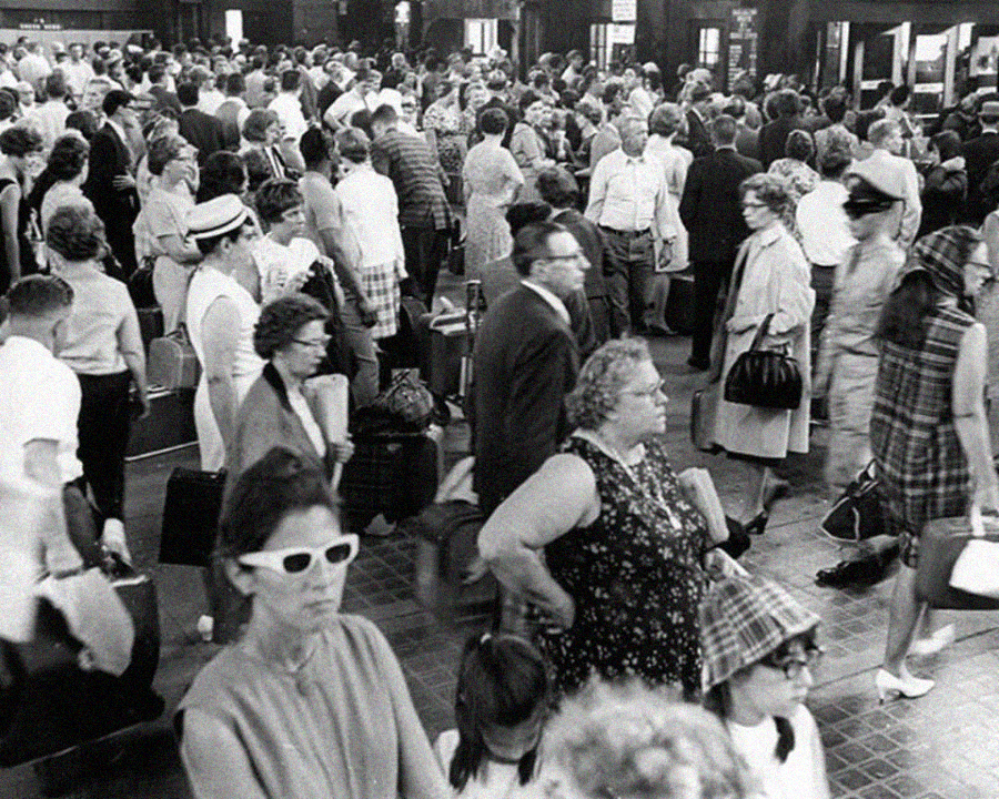 Black and white photo of a large crowd of people, mostly adults, gathered in what appears to be a busy train station. Many are standing, holding bags, and wearing 1960s-style clothing and glasses.