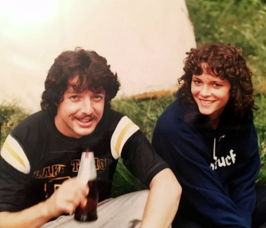 A man with a mustache holding a bottle and a woman with curly hair sit on grass, smiling at the camera. The man wears a black sports jersey, and the woman wears a dark hoodie. A tent is visible in the background.