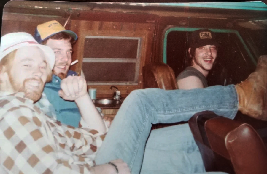 Three men sit in a retro van. Two are in the back, one in plaid and a hat, smiling, and the other wearing a blue jacket and cap, holding a cigarette. The driver is also smiling, wearing a CAT hat, with boots up on the dashboard.