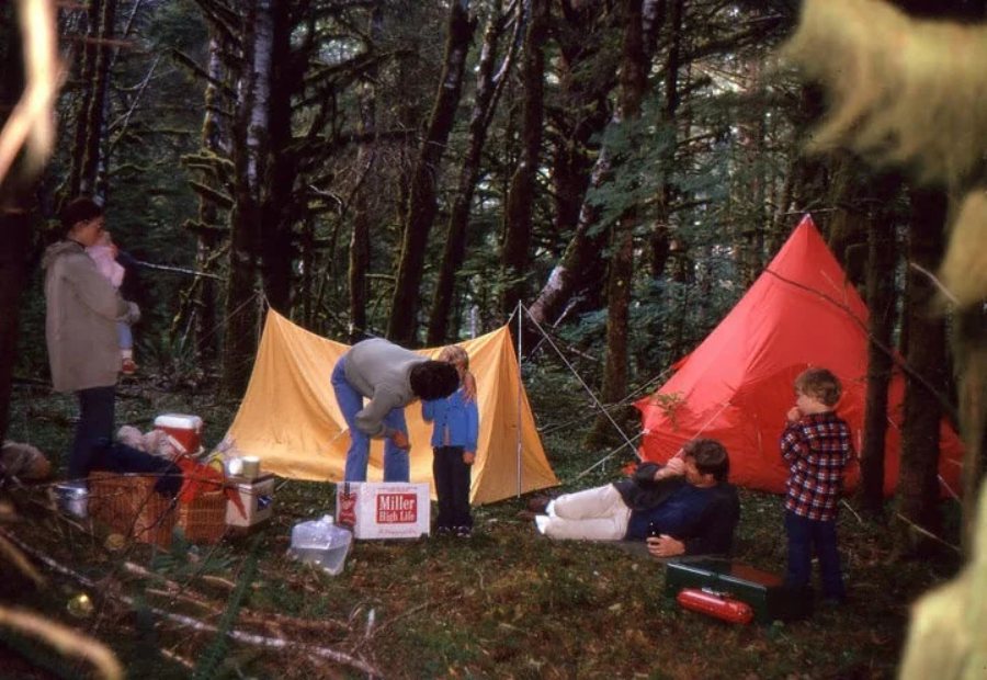 A family camping in a forest with yellow and red tents. Two adults stand near a child, another adult sits on the ground, and two children are nearby. Camping gear and supplies are scattered around.
