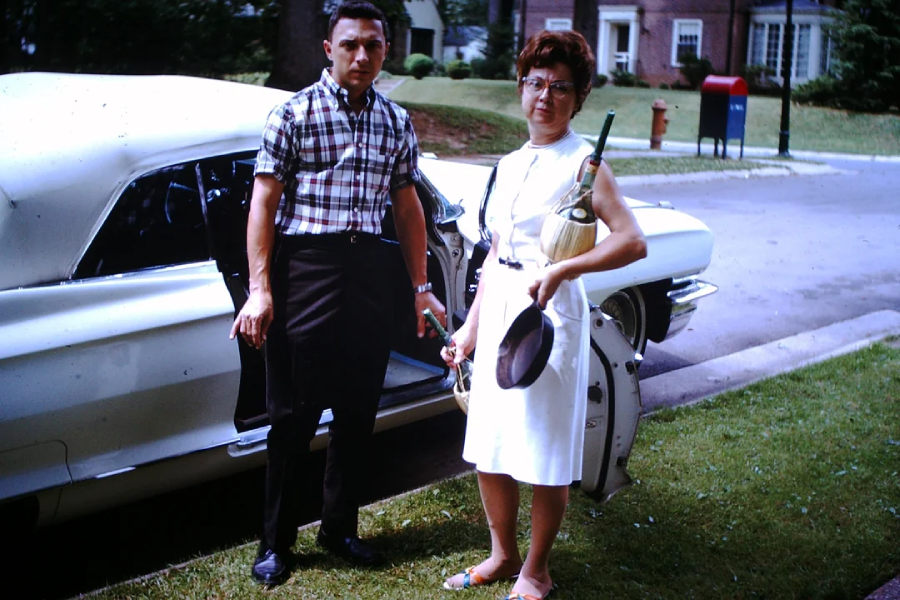 A man in a plaid shirt and dark pants stands next to a woman in a white dress holding cleaning supplies beside a vintage white car parked on a suburban street.