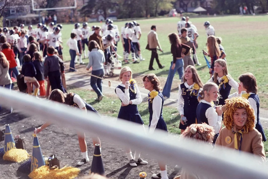 High school cheerleaders in uniforms stand on a sideline, smiling and holding yellow pom-poms, while a football team and spectators gather in the background on a sunny day.