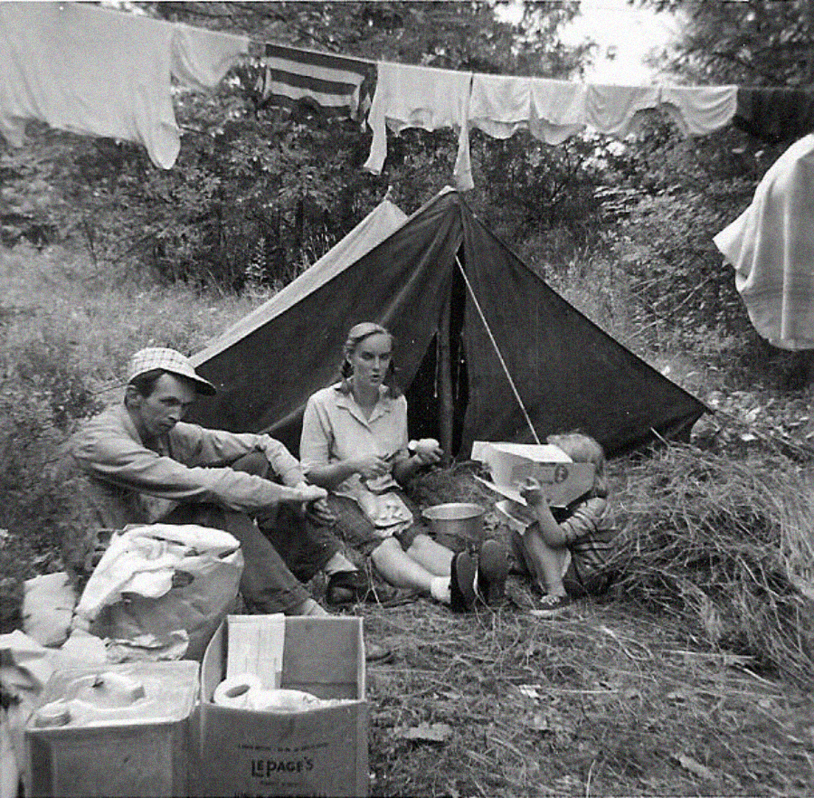A black-and-white photo shows three people sitting on grass by a small tent. A man and two children are near boxes and cooking items, with laundry hanging on a line behind them amid trees and foliage.