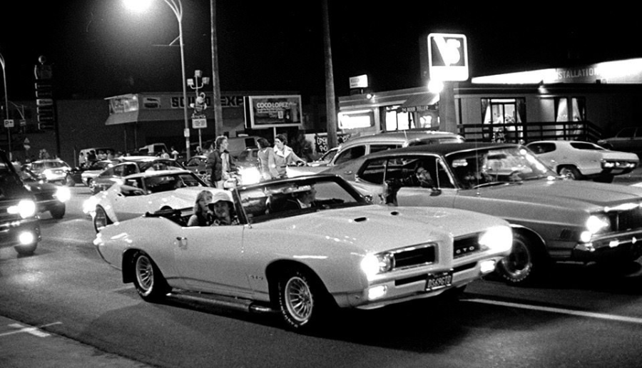 A black and white photo of classic cars cruising on a busy street at night, with people driving and gathering outside illuminated storefronts and diners.