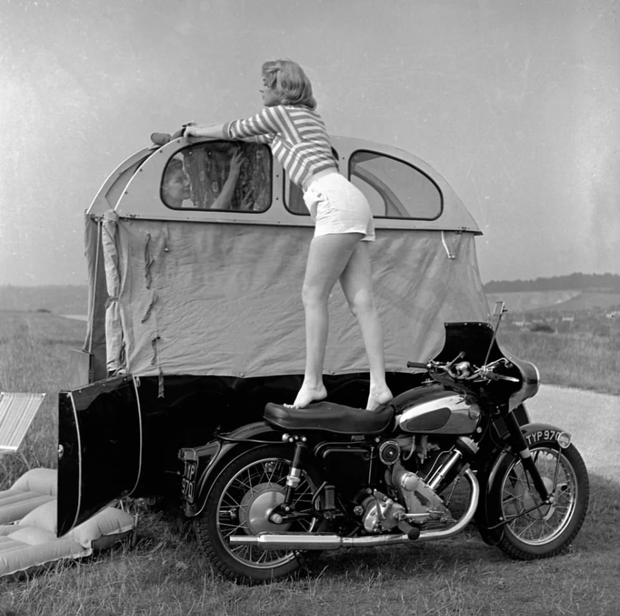 A woman in shorts and a striped top stands on a motorcycle seat to reach and adjust the window of a small, vintage trailer tent set up in an open grassy field.