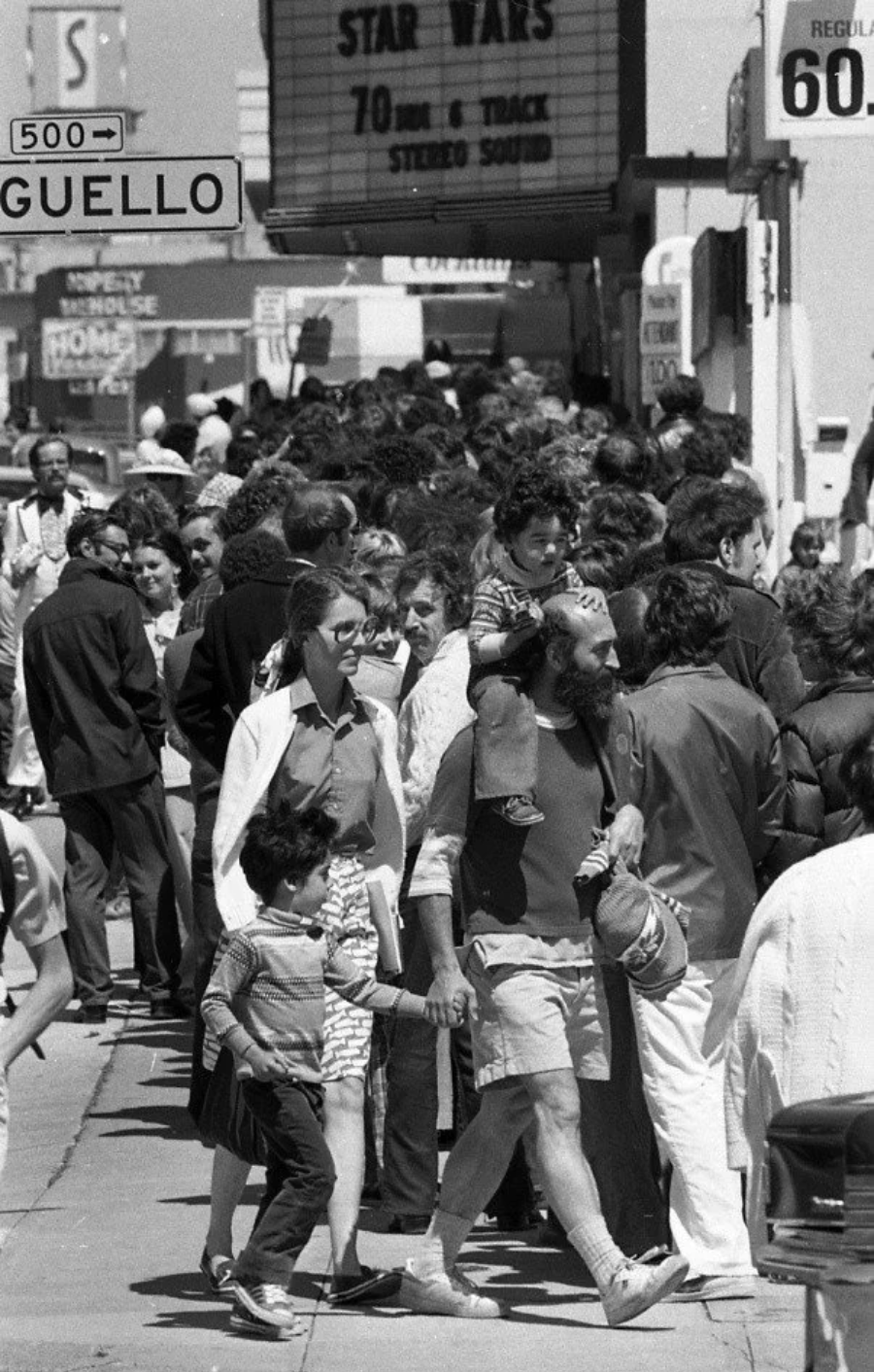 A large crowd lines up outside a theater showing "Star Wars," while families and individuals walk along the busy sidewalk on a sunny day. A street sign with “GUELLO” is visible.
