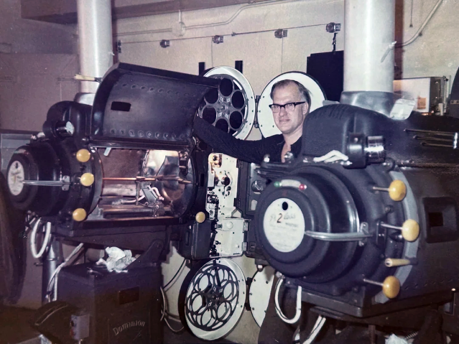 A man stands beside large vintage film projectors in a projection room, with film reels visible on the equipment and behind him. He is wearing glasses and appears to be operating or inspecting the machinery.