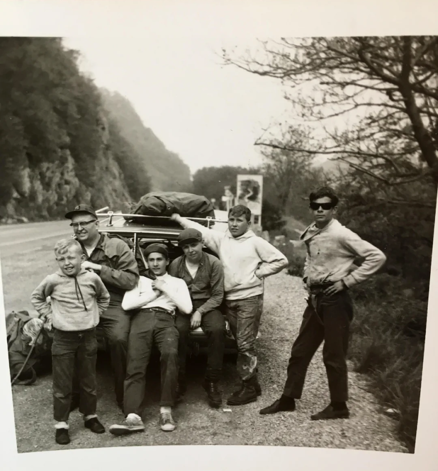 A group of six people, including children and adults, pose by a car loaded with luggage on a roadside, with trees and hills in the background. Some lean on the car while others stand nearby.