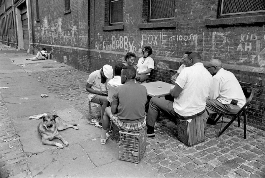 A group of people sit around a table on a cobblestone sidewalk by a brick wall with chalk graffiti, playing a game. A dog lies nearby, and a person sits alone in the background.