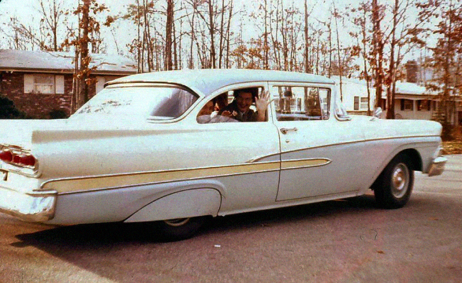 A person waves from the driver’s seat of a vintage light-colored car parked on a suburban street, with houses and leafless trees visible in the background.