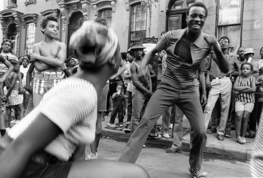 A group of people gathers on a city sidewalk, smiling and watching two people energetically dancing in the foreground. Brownstone buildings are in the background, and the crowd includes children and adults.