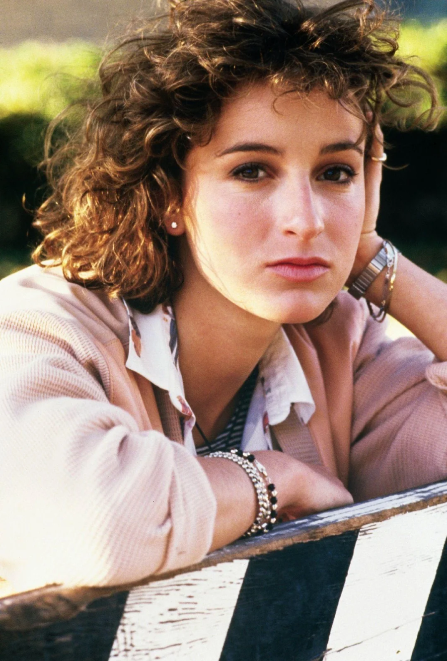 A young woman with curly brown hair rests her head on her hand, looking thoughtfully at the camera. She is wearing a light pink jacket, a patterned white shirt, and several bracelets. A blurred green background is visible.