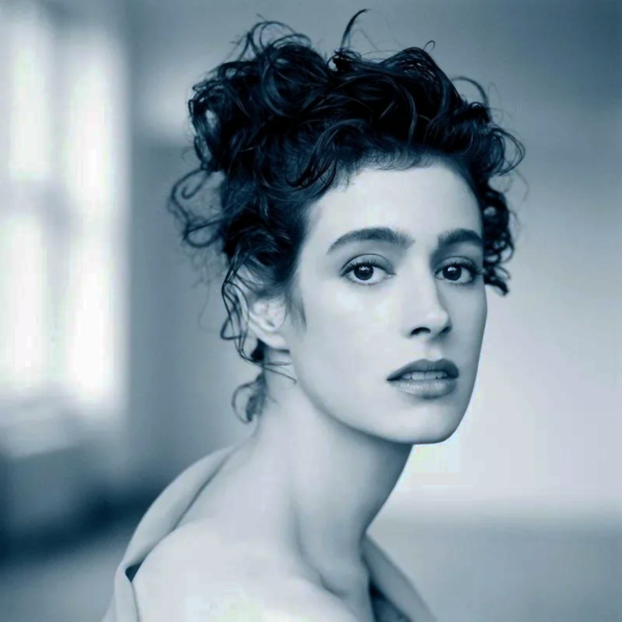A woman with curly dark hair styled up gazes softly at the camera. She has delicate features and wears a bare-shouldered top. The photo is in cool, muted tones with a blurred background.