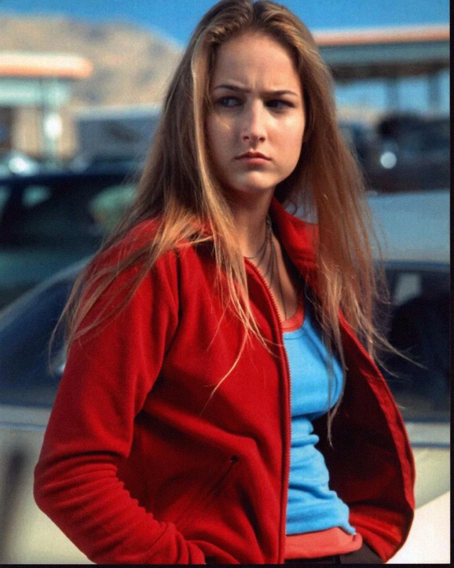 A young woman with long blonde hair stands outdoors in front of a car, wearing a red jacket over a blue top. She looks to the side with a serious expression, with a blurred background of buildings and mountains.