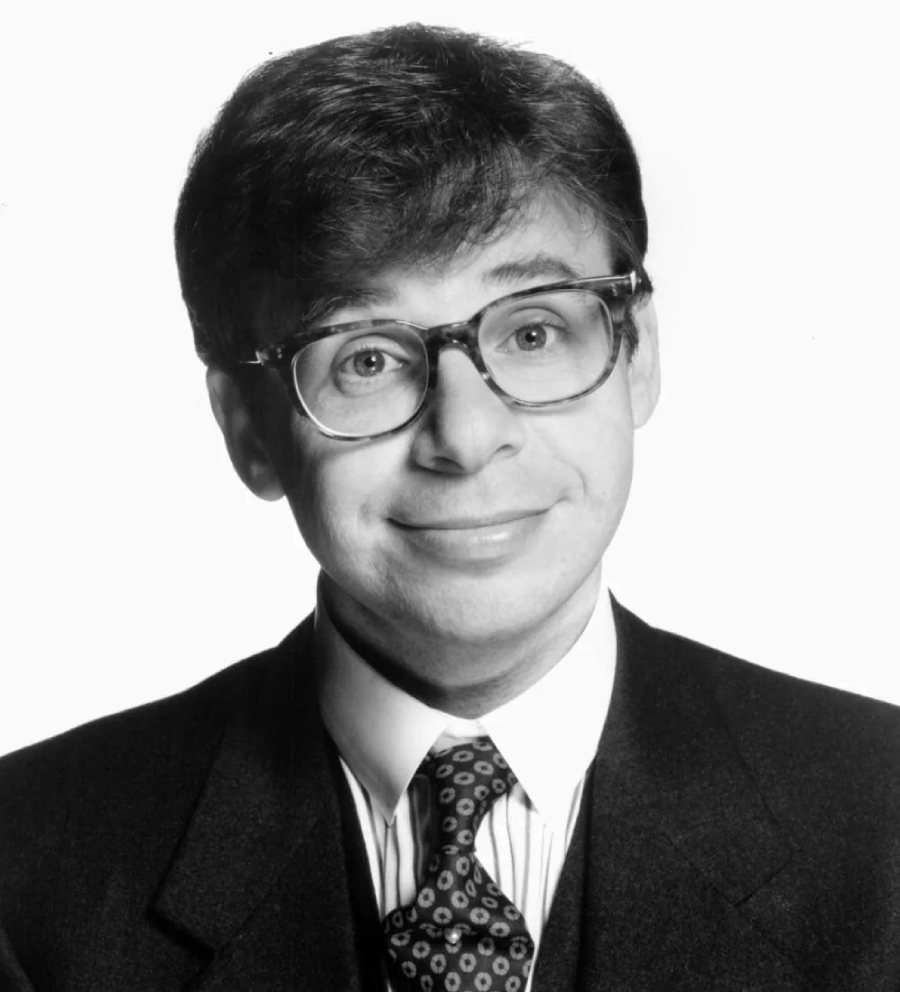 A man with short, dark hair and large glasses is smiling slightly. He is wearing a suit, patterned tie, and collared shirt. The photo is in black and white, with a plain white background.