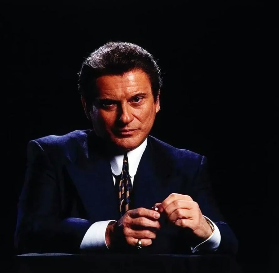 A man in a dark suit and tie sits against a black background, with his hands clasped on a table in front of him. His expression is serious, and he gazes directly at the camera.