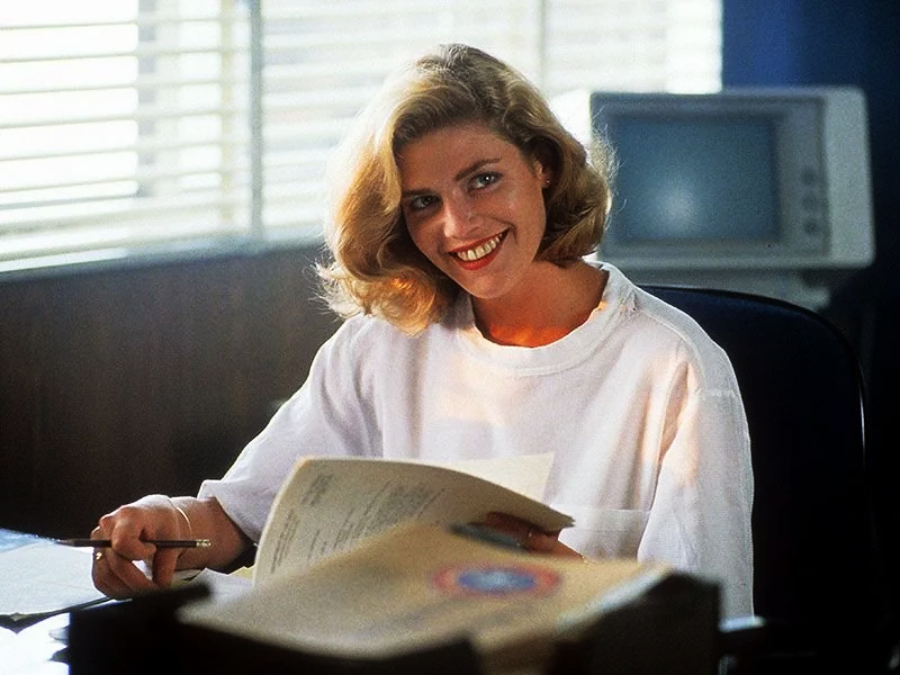 A woman with blonde hair and a white shirt sits at a desk, smiling, holding a pen and looking up from paperwork. Behind her, there is a computer monitor and window blinds letting in natural light.