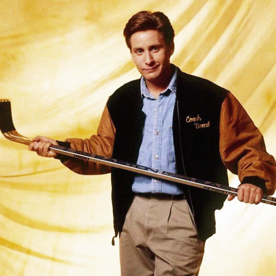 A man wearing a black and brown jacket that says "Coach Bombay," a light blue shirt, and khaki pants holds a hockey stick in both hands against a yellow, softly lit background.