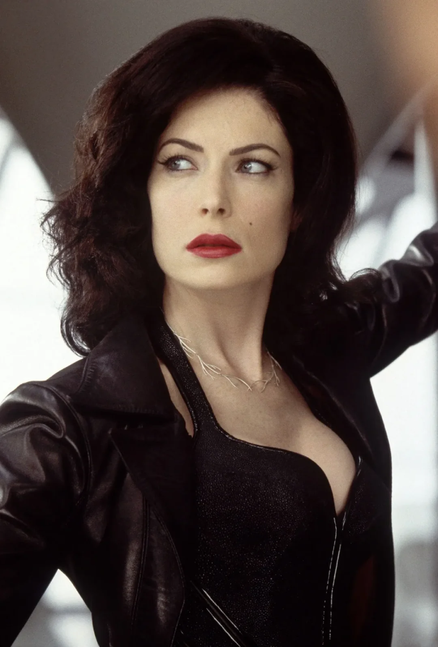 A woman with wavy dark hair, red lipstick, and a black leather jacket looks to the side with a serious expression in a dramatic, softly lit setting.