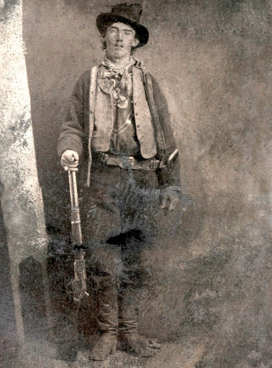 A black-and-white old photograph of a young man in cowboy attire, wearing a hat and jacket, standing and resting his hand on a rifle. The image has a vintage, worn appearance.
