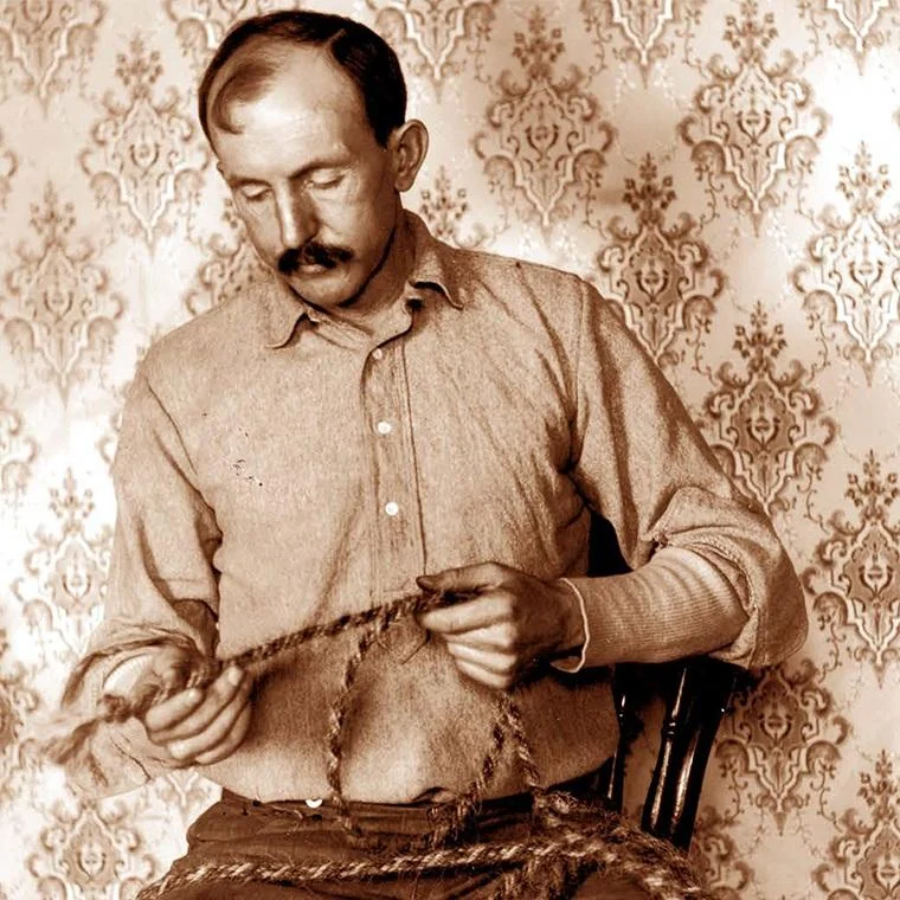 A sepia-toned photo of a man with a mustache sitting on a chair, looking down as he braids or works with thick rope, against a patterned wallpaper background.