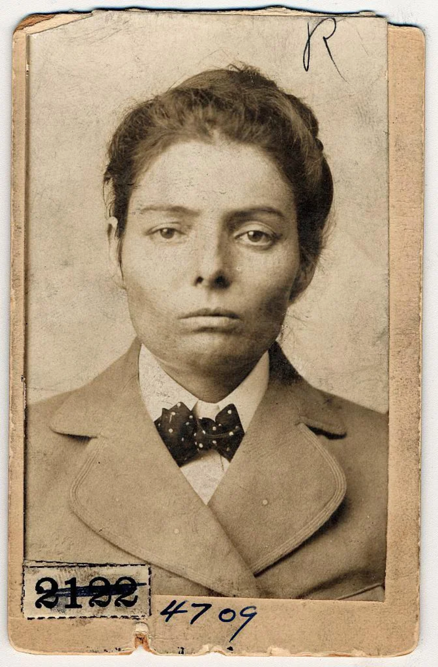 A sepia-toned vintage mugshot of a woman with dark hair tied back, wearing a suit and bow tie. The photo is aged, with handwritten marks and numbers at the bottom and top corners.