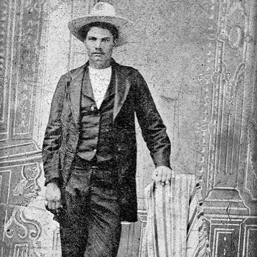 Black and white vintage photograph of a man with a mustache wearing a suit and wide-brimmed hat, standing beside a striped chair in front of an ornate studio backdrop.