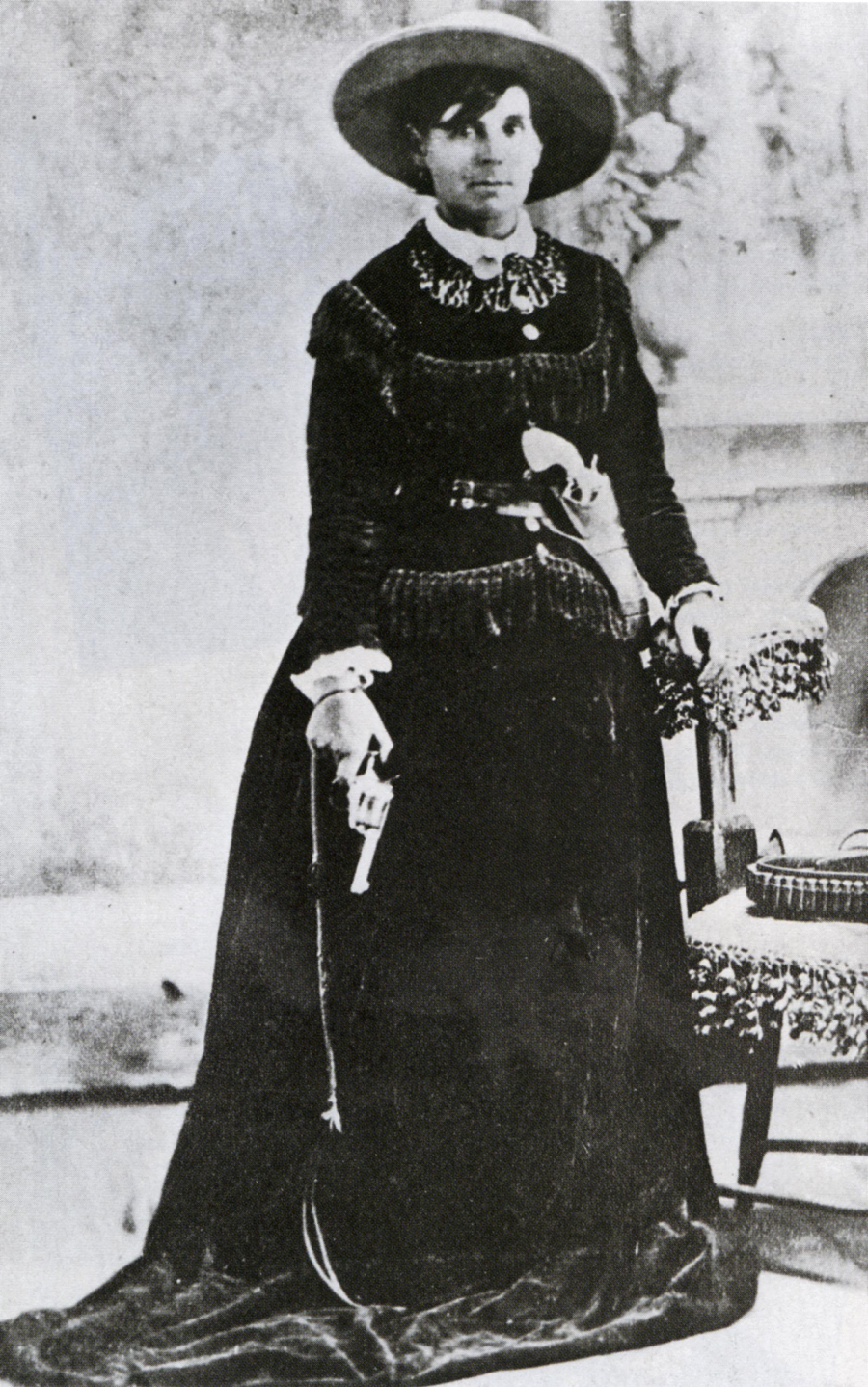 A woman in a long dark dress and wide-brimmed hat stands beside a chair, holding a revolver in her right hand, with a serious expression in a vintage, black-and-white photograph.