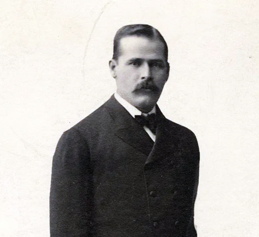 A man with a mustache wearing a dark suit and bow tie stands facing the camera against a plain, light background.