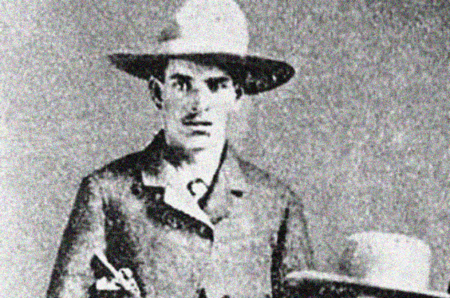 A black-and-white vintage photograph of a man wearing a wide-brimmed hat and a suit jacket, staring directly at the camera with a serious expression. A second, lighter-colored hat rests on a surface beside him.