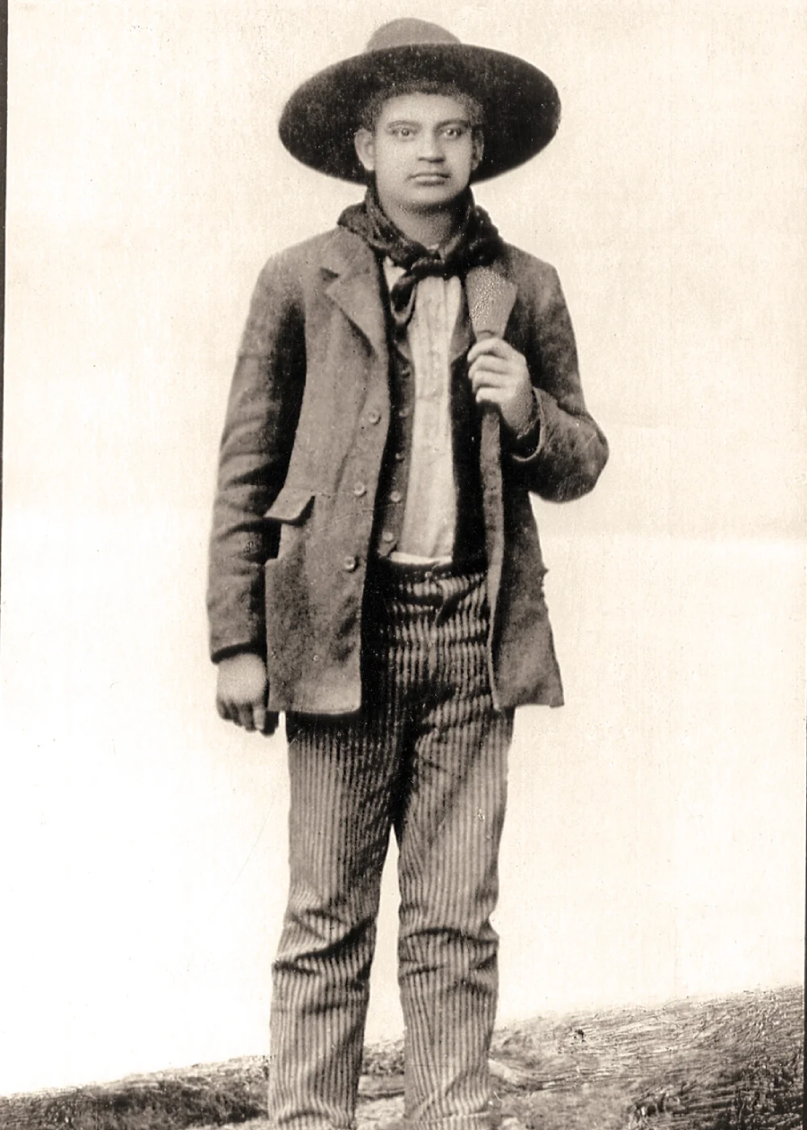 A young person stands facing the camera, wearing a wide-brimmed hat, jacket, neckerchief, striped pants, and boots, with one hand holding their jacket lapel. The photo appears old and sepia-toned.