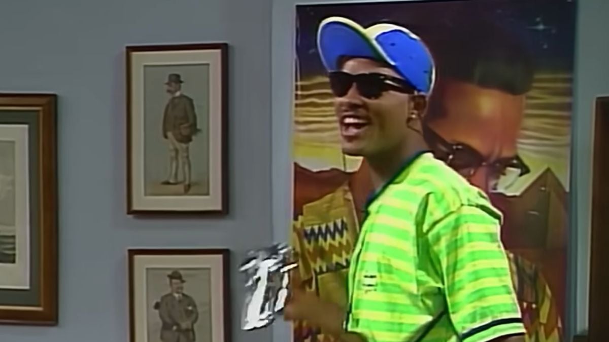A man in a neon green striped shirt, blue and green cap, and sunglasses smiles in a room with framed art and a colorful portrait in the background.