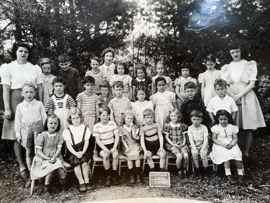 A black-and-white photo of a children's school group, with about 28 young kids and two female adults, posing outdoors among trees. A sign in front reads "Campomila School, Grade 1, Keremeos.
