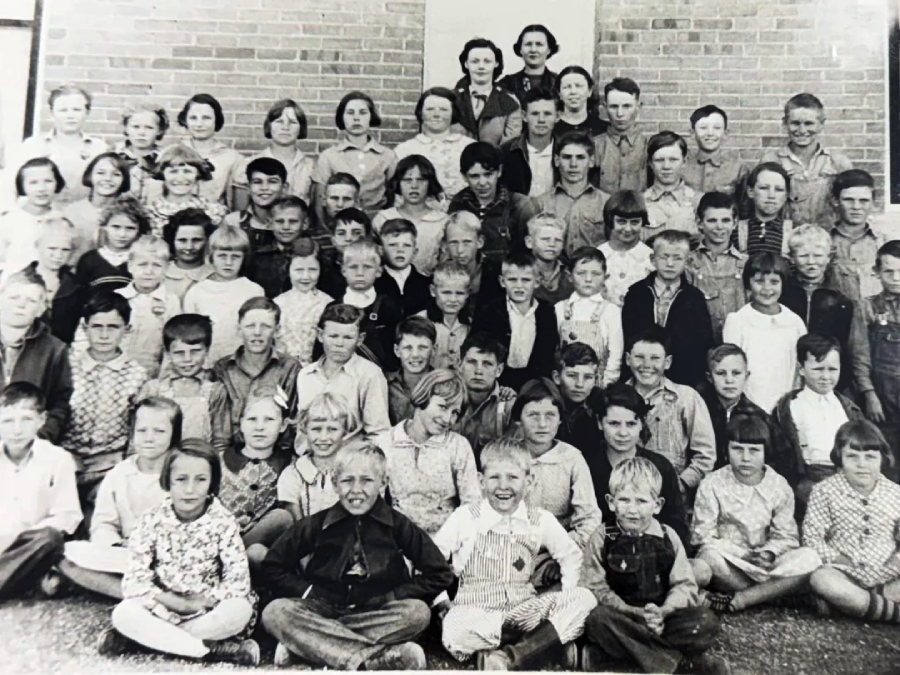 A large group of children and two adults pose for a black-and-white photo in front of a brick building. The children are arranged in several rows, sitting and standing, and appear to be from an earlier era.