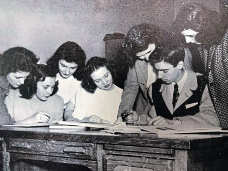A black-and-white photo of six young women and one young man gathered around a desk, looking at papers and writing, possibly working together on a group project.