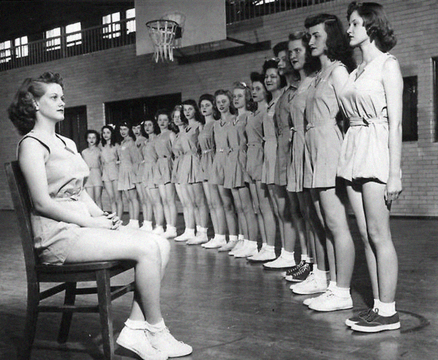 A group of young women in athletic uniforms stand in a line in a gymnasium, facing a seated woman who appears to be their coach or instructor, with a basketball hoop visible above them.