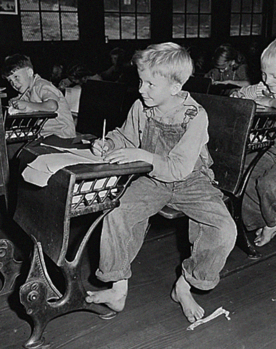 A young boy in overalls sits barefoot at an old-fashioned school desk, writing on paper with a pencil in a classroom. Other children are visible in the background, also seated at desks.