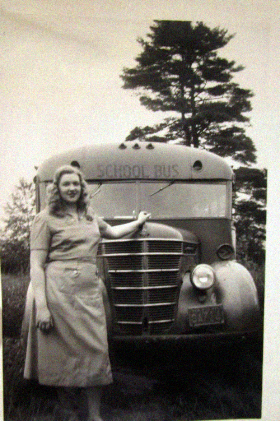A woman in a light dress stands smiling with one arm resting on an old-fashioned school bus, with trees and grass in the background. The bus has "SCHOOL BUS" written above the windshield.