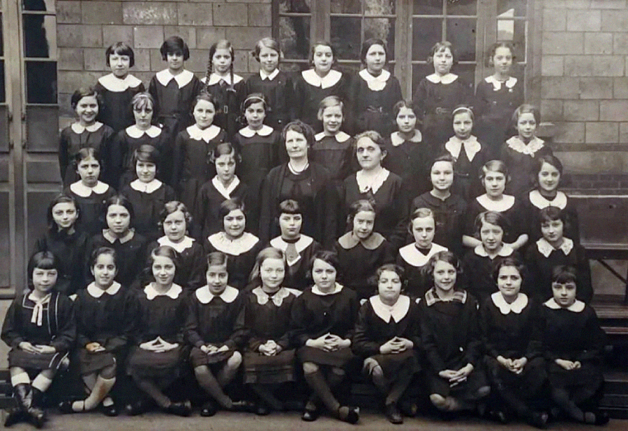 A black-and-white photo of a school class, showing several rows of young girls in dark dresses with white collars, seated and standing, with two adult women in the center, in front of a brick building.