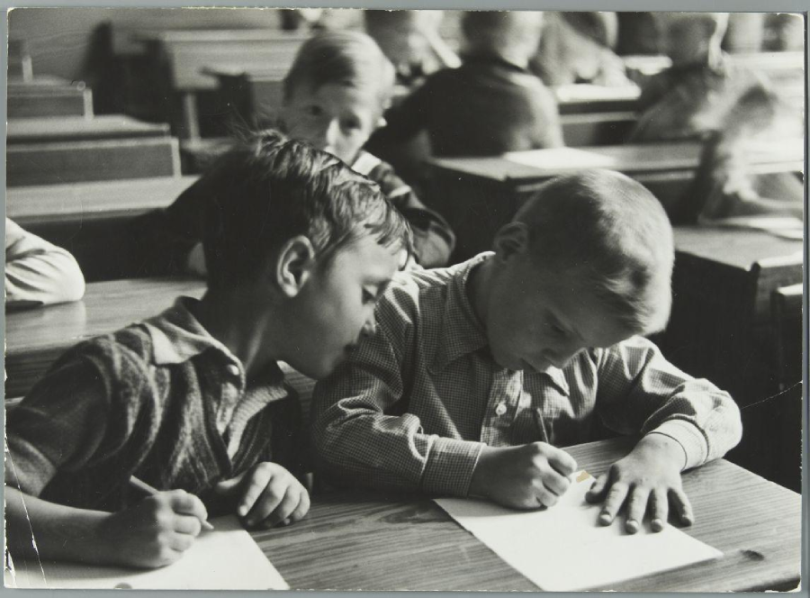 Two young boys sit at a desk in a classroom. One boy appears to be copying or looking at the other's writing. Several other children are seated at desks in the blurred background. The image is black and white.