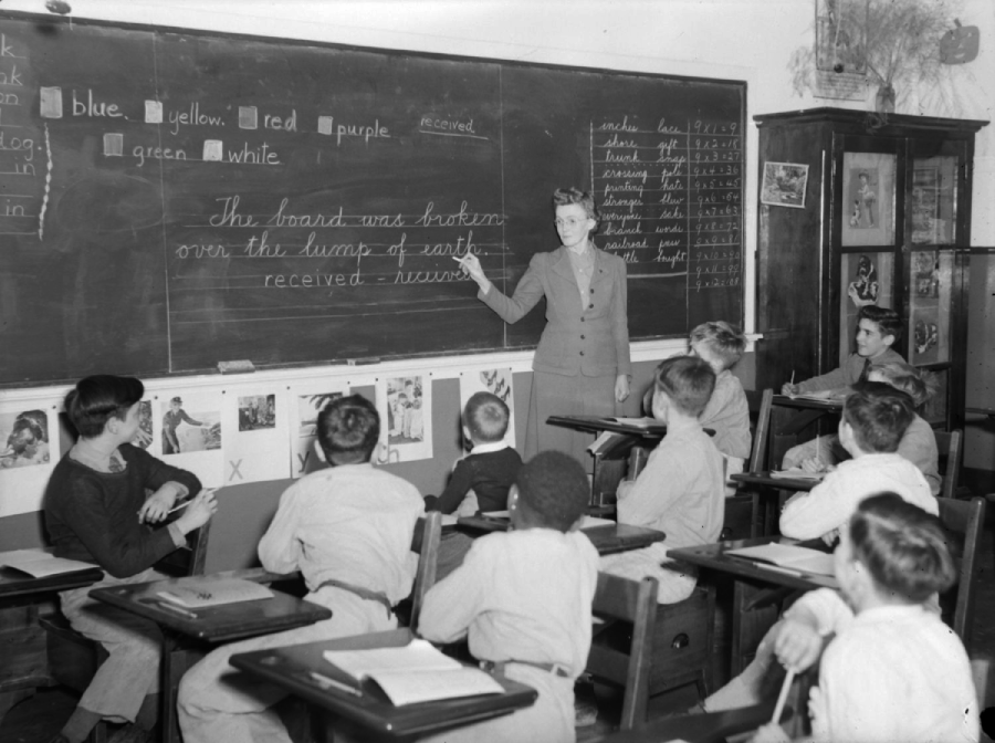 A teacher stands at a chalkboard pointing to cursive writing, while several students seated at desks watch and take notes in a classroom filled with educational charts and posters.