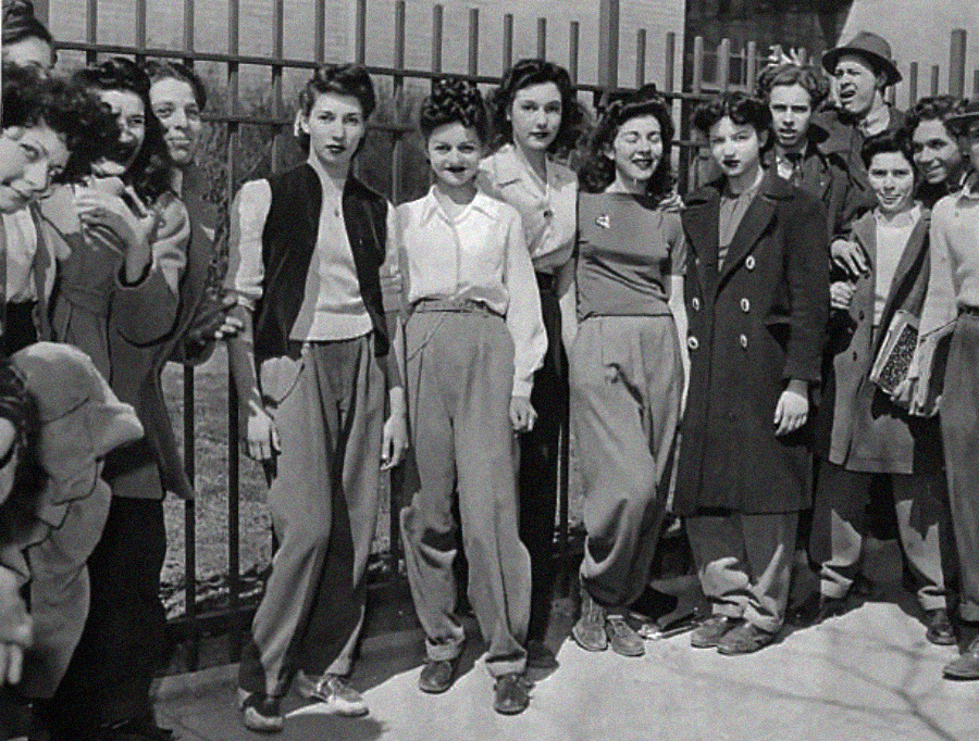 A group of young women and men stand and smile near a metal fence, dressed in mid-20th-century clothing, including high-waisted pants, jackets, and coats. They appear to be socializing outdoors in a city setting.