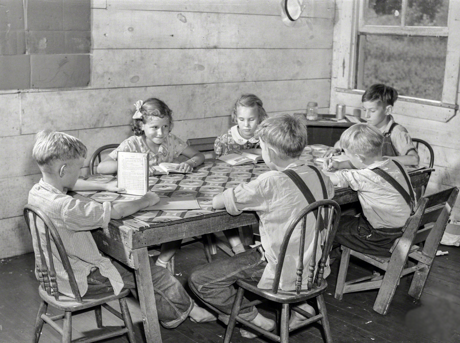 Six young children sit around a wooden table in a classroom, reading from books. The room has wooden walls and a window, and the children appear focused on their work.
