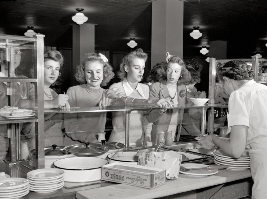 Four women stand in line at a cafeteria counter, looking at the food options while a server prepares dishes. The women have 1940s-style hair and clothing, and various expressions of anticipation or curiosity.