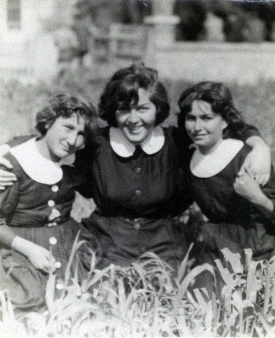 Three young women wearing dark dresses with white collars sit close together outdoors, smiling and embracing each other, surrounded by tall grass with blurred trees in the background. The photo appears to be old and black and white.